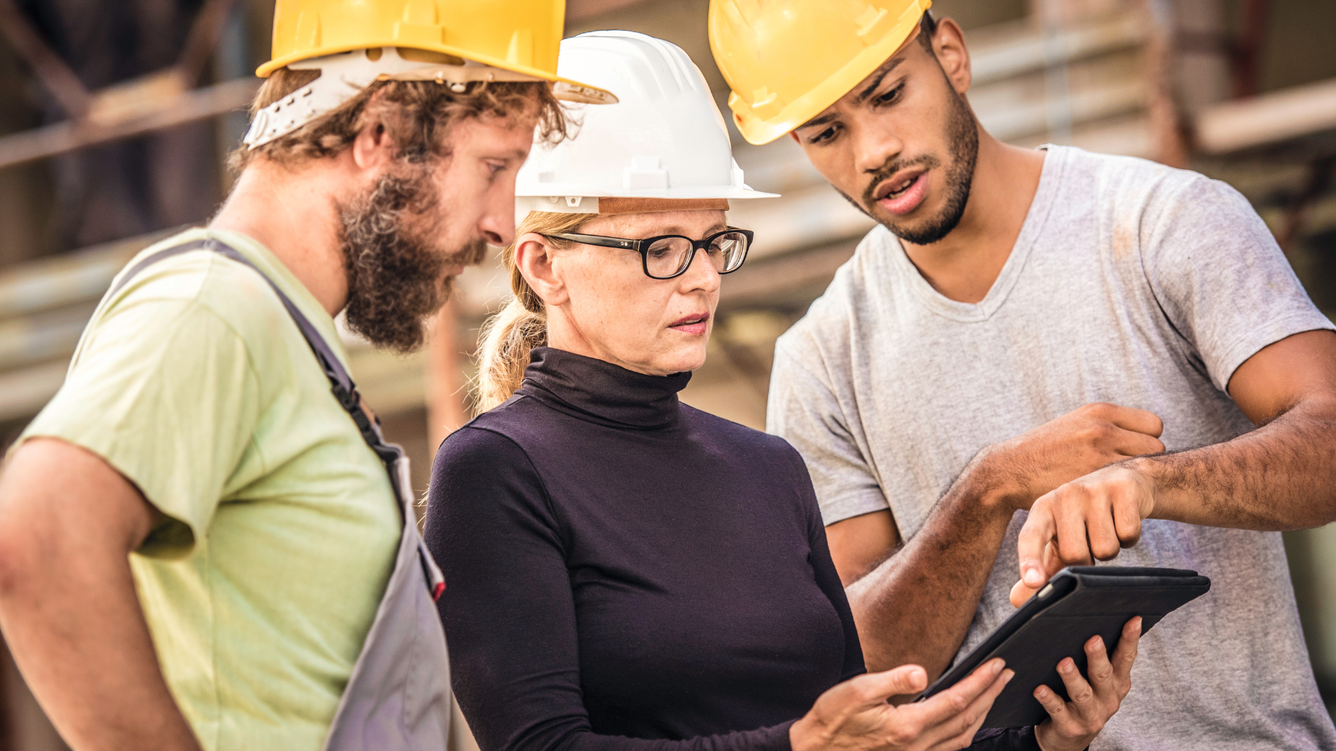 Three people at a construction site wearing hard hats, reviewing a tablet