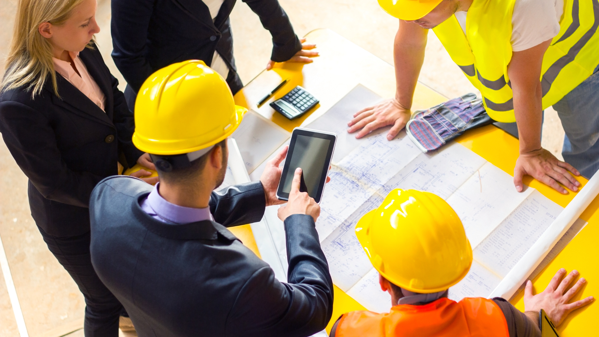 People in suits and people in construction attire and hard hats working together. Looking at blueprints and tablets.